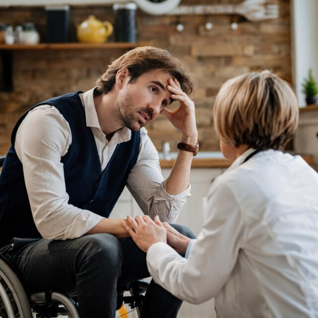young worried man wheelchair communicating with doctor while she is visiting him home